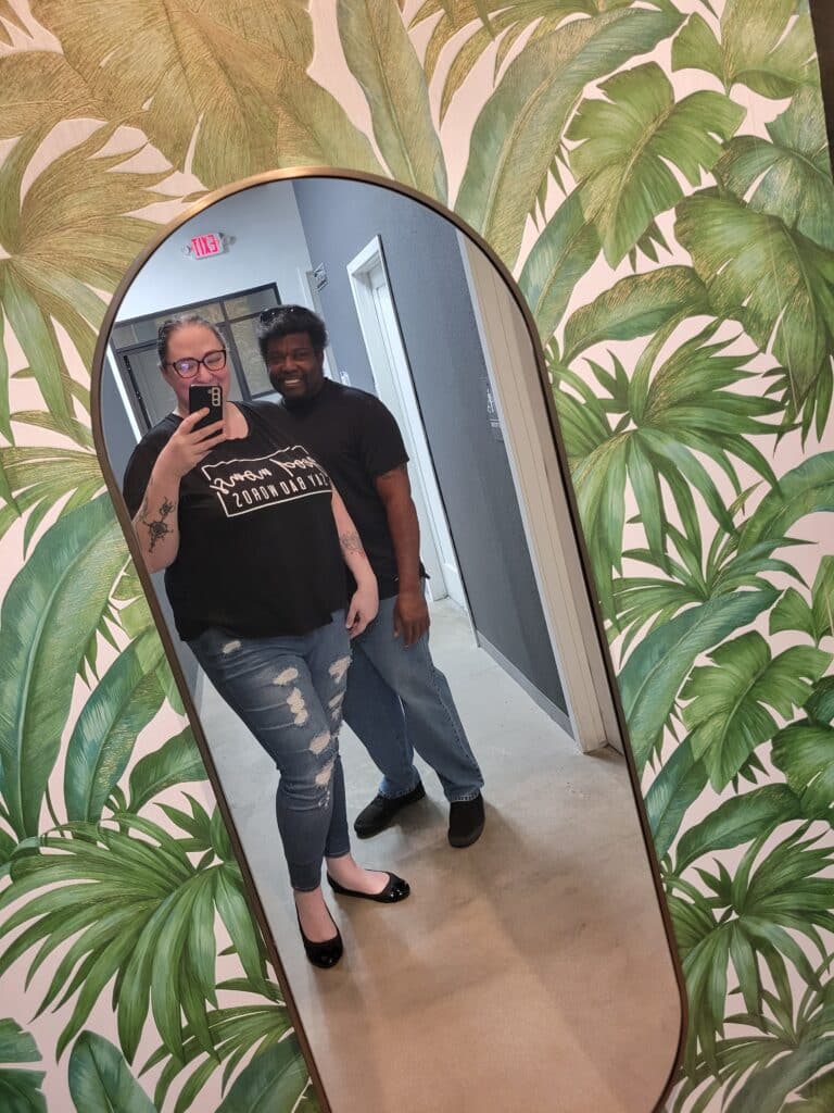 Sadie standing on the left in a black shirt and jeans, Kevin on the left in a black shirt and jeans, they are looking in a mirror. There are green palm trees on the wall paper.